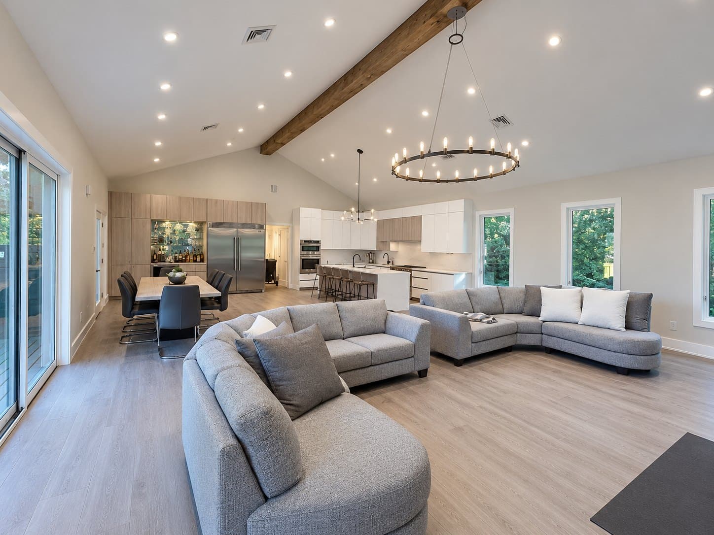 An open-plan living room under a vaulted ceiling with a ring chandelier, kitchen beyond